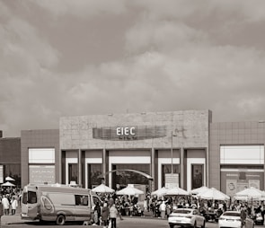 A large building with signage reading 'EIEC' is visible, surrounded by a crowd of people gathered in front of it. Vehicles, including a van and a car, are parked nearby. Several umbrellas are set up outside the building, possibly for outdoor seating or shelter.