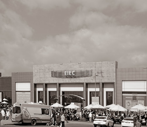 A large building with signage reading 'EIEC' is visible, surrounded by a crowd of people gathered in front of it. Vehicles, including a van and a car, are parked nearby. Several umbrellas are set up outside the building, possibly for outdoor seating or shelter.