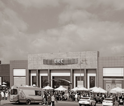 A large building with signage reading 'EIEC' is visible, surrounded by a crowd of people gathered in front of it. Vehicles, including a van and a car, are parked nearby. Several umbrellas are set up outside the building, possibly for outdoor seating or shelter.