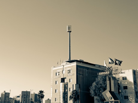A tall building with a prominent vertical signpost on its roof extends into the sky. The building features modern architecture with a combination of glass and brickwork. Several flags are flying on the rooftop, and a sign reads 'Mansoura College' at the front. Surrounding the building are a few palm trees and other buildings in the background under a clear sky.