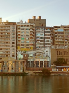 A row of densely packed residential buildings by a riverbank, showcasing a variety of architectural styles. The buildings exhibit a mix of balconies, windows, and air conditioning units. Some signs with Arabic script are visible, and the scene captures the evening light casting a warm hue over the facade.