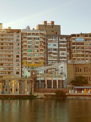 A row of densely packed residential buildings by a riverbank, showcasing a variety of architectural styles. The buildings exhibit a mix of balconies, windows, and air conditioning units. Some signs with Arabic script are visible, and the scene captures the evening light casting a warm hue over the facade.