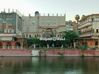 A waterfront scene featuring a collection of buildings with ornate architectural details. There's a prominent sign in Arabic positioned above a lush green tree, surrounded by various types of building facades. The river reflects these structures, and palm trees are visible near the water's edge.