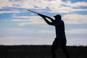 A silhouette of a person aiming a shotgun in an open field, with a background of a partly cloudy sky. The individual is wearing a cap and appears to be engaged in an outdoor activity, possibly hunting.