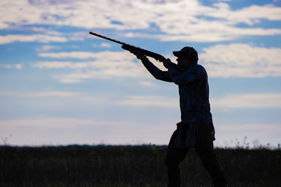 A silhouette of a person aiming a shotgun in an open field, with a background of a partly cloudy sky. The individual is wearing a cap and appears to be engaged in an outdoor activity, possibly hunting.