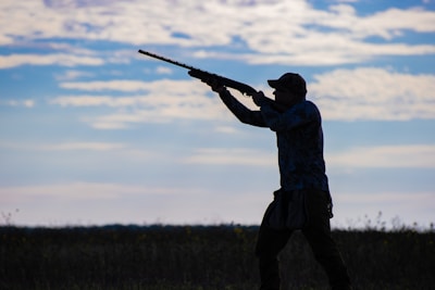 A silhouette of a person aiming a shotgun in an open field, with a background of a partly cloudy sky. The individual is wearing a cap and appears to be engaged in an outdoor activity, possibly hunting.