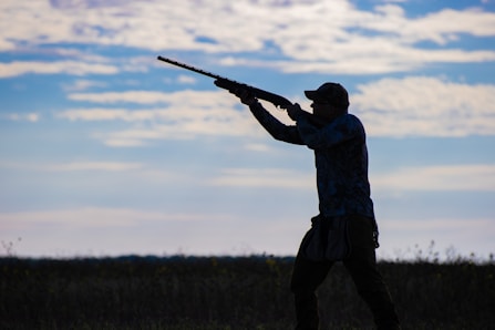 A silhouette of a person aiming a shotgun in an open field, with a background of a partly cloudy sky. The individual is wearing a cap and appears to be engaged in an outdoor activity, possibly hunting.