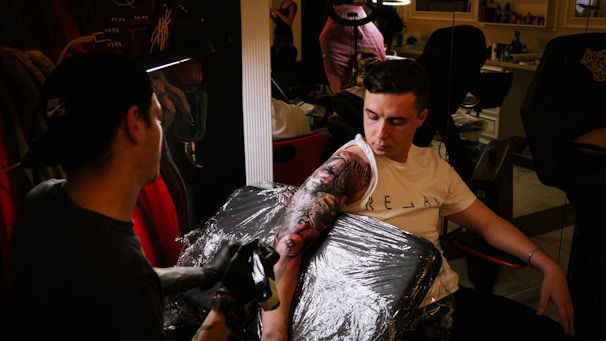 A tattoo artist is applying a detailed black and grey tattoo to a person's arm. The person receiving the tattoo is seated in a black chair covered with clear plastic wrap. The artist wears a cap and gloves while concentrating on their work. The setting appears to be a tattoo parlor with visible mirrors, chairs, and various tattooing equipment in the background.