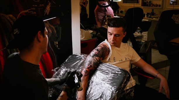 A tattoo artist is applying a detailed black and grey tattoo to a person's arm. The person receiving the tattoo is seated in a black chair covered with clear plastic wrap. The artist wears a cap and gloves while concentrating on their work. The setting appears to be a tattoo parlor with visible mirrors, chairs, and various tattooing equipment in the background.