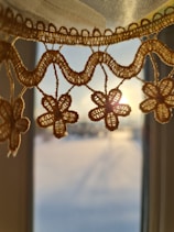 Intricate crochet lace with floral patterns is highlighted by the warm glow of sunlight streaming through a window. The lace hangs delicately, casting soft shadows on the blurred backdrop of a serene outdoor scene.