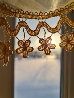Intricate crochet lace with floral patterns is highlighted by the warm glow of sunlight streaming through a window. The lace hangs delicately, casting soft shadows on the blurred backdrop of a serene outdoor scene.