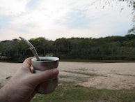 A hand holding a metal cup containing a drink with a metal straw, against a backdrop of a sandy area, a river, and a dense forest under a partly cloudy sky.