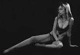 A woman sits in a somewhat relaxed pose against a dark background, wearing black lace underwear. Her expression is contemplative as she looks off to the side. The image is in black and white, highlighting the contrast and shadows.
