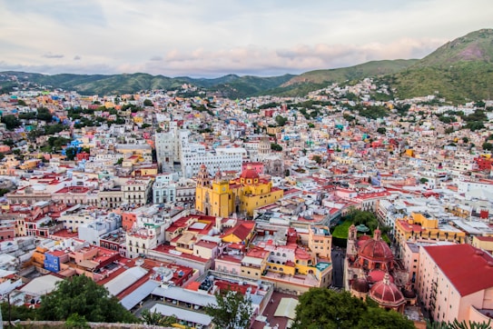 An aerial view of a colorful cityscape nestled among green hills. The buildings are painted in a variety of vibrant hues, including yellow, red, blue, and pink, creating a lively and visually striking scene. The architecture appears historical, with some prominent church towers and a mix of colonial style buildings. The layout of the city is dense, with narrow streets winding between the closely packed structures.
