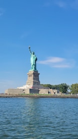 The image features the Statue of Liberty on Liberty Island with a clear blue sky in the background. The statue stands on a stone pedestal, with the water of New York Harbor in the foreground.