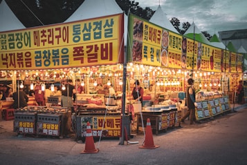A vibrant street market scene with brightly lit food stalls adorned with colorful signs in Korean script. Several vendors and customers can be seen interacting around the stalls, which display various types of food, cooking equipment, and ingredients. The atmosphere is lively, with warm hanging lights creating an inviting ambience in the evening setting.