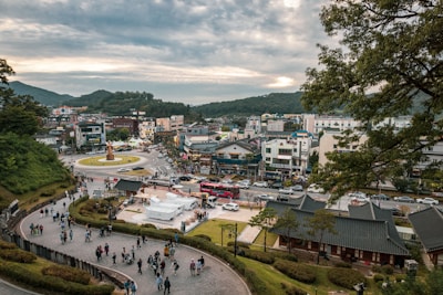 A scenic cityscape featuring a bustling urban area surrounded by lush green hills. In the foreground, a diverse group of people walks along a paved pathway that curves through a park. Traditional buildings with sloped roofs are visible, adjacent to modern structures. At the center is a roundabout with a statue, and several cars and buses navigate through the streets.
