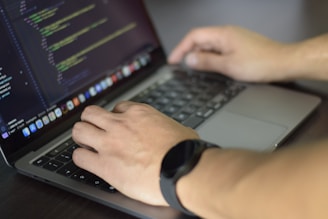 A person is typing code on a laptop. The screen displays a code editor with lines of code visible. The individual is wearing a watch on their left wrist and their hands are positioned over the keyboard.