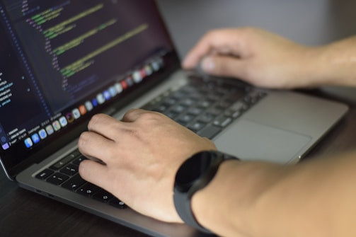 A person is typing code on a laptop. The screen displays a code editor with lines of code visible. The individual is wearing a watch on their left wrist and their hands are positioned over the keyboard.