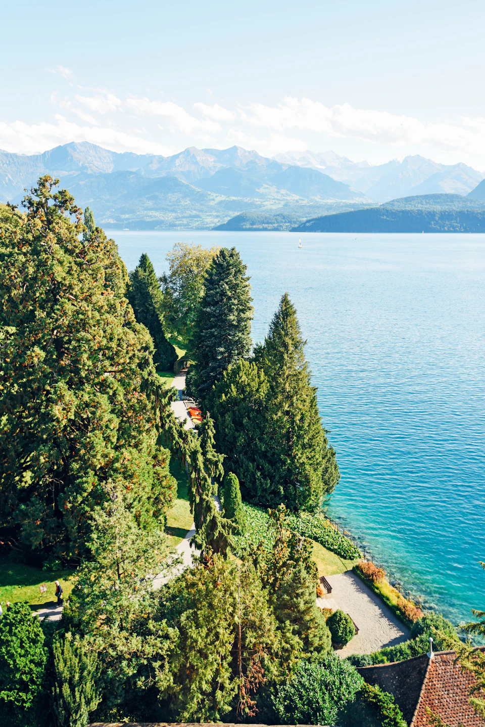 The image depicts a serene landscape featuring a large lake surrounded by lush greenery, including trees and bushes. The calm water reflects the clear blue sky above, creating a tranquil atmosphere. In the foreground, there is a path leading towards the lake, inviting visitors to explore its beauty. On one side of the lake, several boats are docked, suggesting recreational activities such as boating or fishing. The image is taken from an elevated perspective, providing a comprehensive view of the landscape and its various elements.