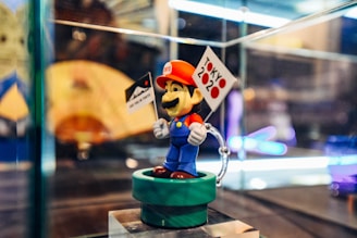 A figurine of a well-known video game character dressed in blue overalls, a red hat, and a red shirt, standing on a green platform. The character is holding two small flags, one with the text 'Tokyo 2020' featuring red dots and another flag with a mountain design. The background is slightly out of focus with colorful elements, possibly representing a display case.