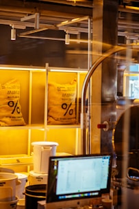 Two large burlap bags labeled 'ARABICA' are placed on illuminated shelves in what appears to be a coffee shop or coffee roasting area. Below the bags, there are white and black plastic containers, and in the foreground, there is a computer monitor displaying some information. Industrial pipes and structures are visible in the background.
