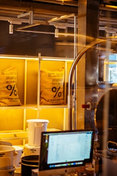 Two large burlap bags labeled 'ARABICA' are placed on illuminated shelves in what appears to be a coffee shop or coffee roasting area. Below the bags, there are white and black plastic containers, and in the foreground, there is a computer monitor displaying some information. Industrial pipes and structures are visible in the background.