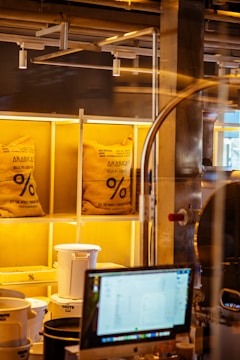Two large burlap bags labeled 'ARABICA' are placed on illuminated shelves in what appears to be a coffee shop or coffee roasting area. Below the bags, there are white and black plastic containers, and in the foreground, there is a computer monitor displaying some information. Industrial pipes and structures are visible in the background.