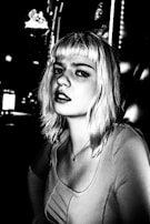 A black and white portrait of a person with light-colored hair, looking directly at the camera. The setting appears to be an urban environment at night, with blurred lights in the background adding a dramatic effect. The person's expression is serious and slightly contemplative. The high contrast enhances the shadows and highlights, giving the image a distinctive and edgy look.