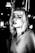 A black and white portrait of a person with light-colored hair, looking directly at the camera. The setting appears to be an urban environment at night, with blurred lights in the background adding a dramatic effect. The person's expression is serious and slightly contemplative. The high contrast enhances the shadows and highlights, giving the image a distinctive and edgy look.