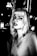 A black and white portrait of a person with light-colored hair, looking directly at the camera. The setting appears to be an urban environment at night, with blurred lights in the background adding a dramatic effect. The person's expression is serious and slightly contemplative. The high contrast enhances the shadows and highlights, giving the image a distinctive and edgy look.