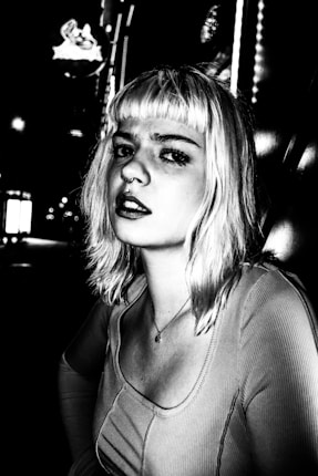 A black and white portrait of a person with light-colored hair, looking directly at the camera. The setting appears to be an urban environment at night, with blurred lights in the background adding a dramatic effect. The person's expression is serious and slightly contemplative. The high contrast enhances the shadows and highlights, giving the image a distinctive and edgy look.