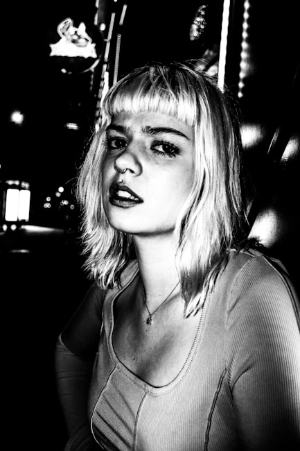 A black and white portrait of a person with light-colored hair, looking directly at the camera. The setting appears to be an urban environment at night, with blurred lights in the background adding a dramatic effect. The person's expression is serious and slightly contemplative. The high contrast enhances the shadows and highlights, giving the image a distinctive and edgy look.