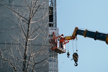Two workers wearing safety gear are in an aerial lift platform attached to a crane, working on a building facade. A leafless tree is in the foreground, and the sky is clear.