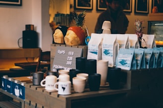 A coffee shop counter displays various items, including cups, mugs, and bags of coffee beans arranged neatly. Two decorative bread loaves wearing beanies are placed on a shelf above, alongside pineapples. The setting has a cozy, dimly lit ambiance with wooden furniture and a menu board showing pricing details.