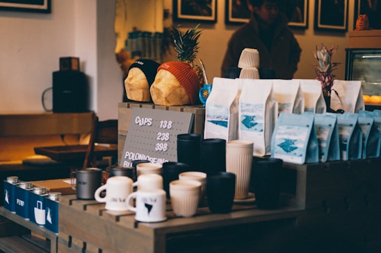 A coffee shop counter displays various items, including cups, mugs, and bags of coffee beans arranged neatly. Two decorative bread loaves wearing beanies are placed on a shelf above, alongside pineapples. The setting has a cozy, dimly lit ambiance with wooden furniture and a menu board showing pricing details.