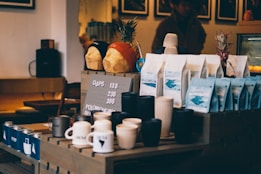 A coffee shop counter displays various items, including cups, mugs, and bags of coffee beans arranged neatly. Two decorative bread loaves wearing beanies are placed on a shelf above, alongside pineapples. The setting has a cozy, dimly lit ambiance with wooden furniture and a menu board showing pricing details.
