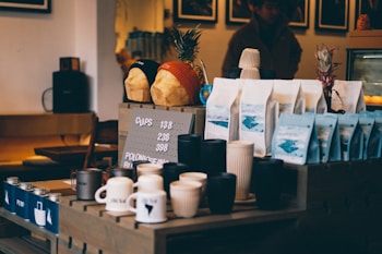 A coffee shop counter displays various items, including cups, mugs, and bags of coffee beans arranged neatly. Two decorative bread loaves wearing beanies are placed on a shelf above, alongside pineapples. The setting has a cozy, dimly lit ambiance with wooden furniture and a menu board showing pricing details.