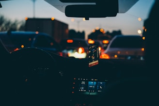 The scene is inside a car during a traffic jam, with a dashboard showing electronic displays and a smartphone mounted, displaying navigation. Headlights from other vehicles create a pattern of red and white lights in the evening ambiance.