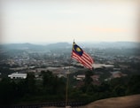 A Malaysian flag is prominently displayed on a pole, overlooking a vast cityscape. Hills and buildings are visible in the distance under a hazy sky.