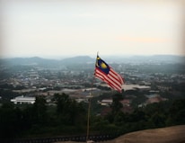 A Malaysian flag is prominently displayed on a pole, overlooking a vast cityscape. Hills and buildings are visible in the distance under a hazy sky.