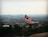 A Malaysian flag is prominently displayed on a pole, overlooking a vast cityscape. Hills and buildings are visible in the distance under a hazy sky.
