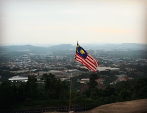 A Malaysian flag is prominently displayed on a pole, overlooking a vast cityscape. Hills and buildings are visible in the distance under a hazy sky.