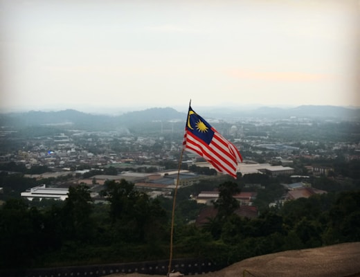 A Malaysian flag is prominently displayed on a pole, overlooking a vast cityscape. Hills and buildings are visible in the distance under a hazy sky.