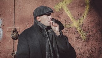 A person wearing a cap, coat, and scarf is smoking a cigarette while standing against a textured red wall with a large yellow hammer and sickle symbol in the background. The person is facing sideways with a serious expression.