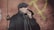 A person wearing a cap, coat, and scarf is smoking a cigarette while standing against a textured red wall with a large yellow hammer and sickle symbol in the background. The person is facing sideways with a serious expression.