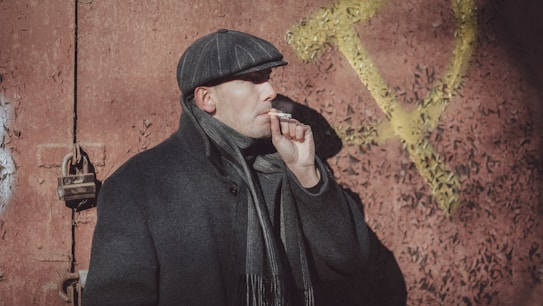 A person wearing a cap, coat, and scarf is smoking a cigarette while standing against a textured red wall with a large yellow hammer and sickle symbol in the background. The person is facing sideways with a serious expression.