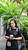 A person wearing a dark apron holds a drink beneath lush green tropical leaves. The background features potted plants and a pathway, suggesting a garden or outdoor setting. The person smiles gently, creating a welcoming atmosphere.