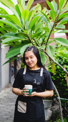 A person wearing a dark apron holds a drink beneath lush green tropical leaves. The background features potted plants and a pathway, suggesting a garden or outdoor setting. The person smiles gently, creating a welcoming atmosphere.