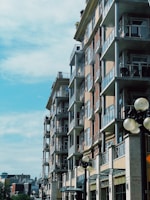 A modern multi-story apartment building with glass balconies under a clear blue sky. The building is made of brick and concrete, with lamp posts visible in the foreground.